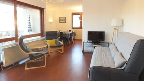 A living room with a sofa, a TV, a dining table with chairs, and a window with a view of a building. - Collioure Town with Parking – Apartment Sorra (Collioure)
