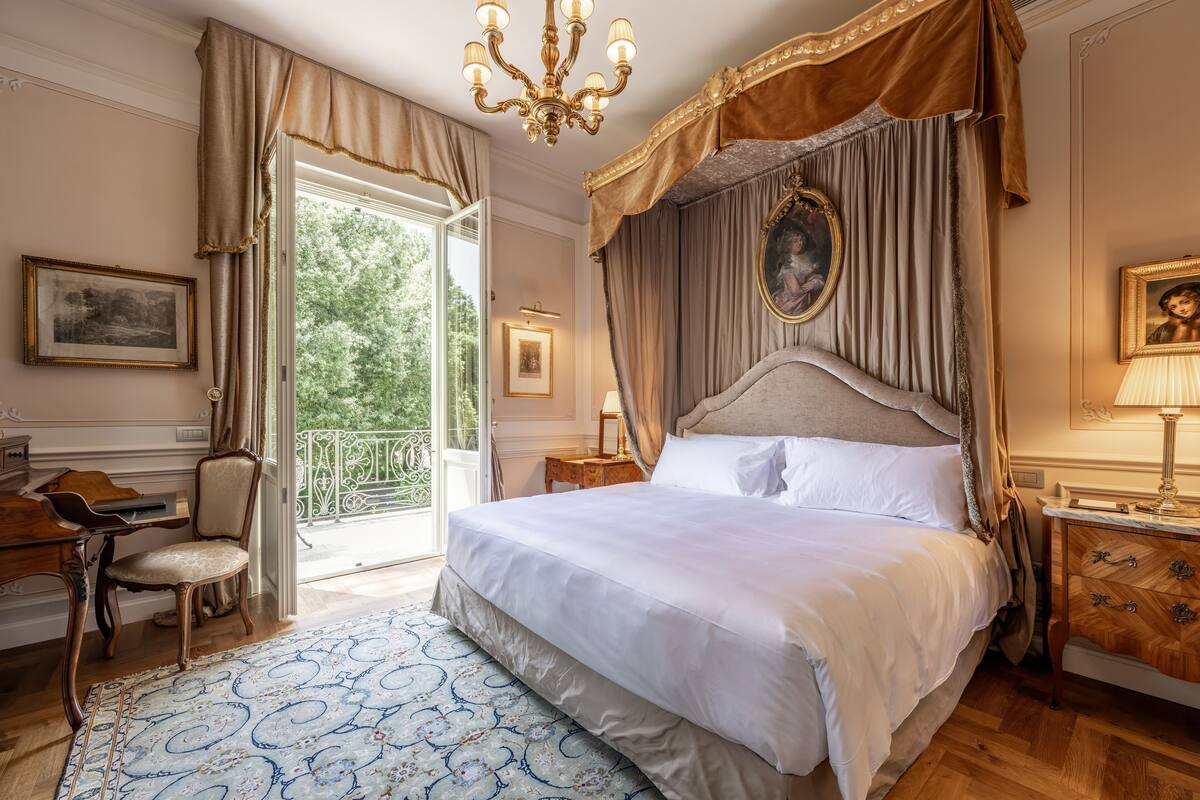 Een slaapkamer met een groot bed, een bureau en een stoel. Er is een balkon met uitzicht op bomen.