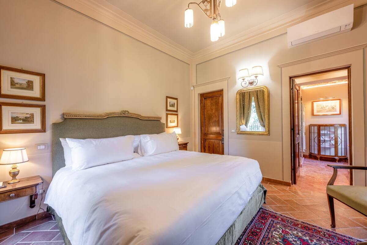 Een goed verlichte hotelkamer met een groot bed, een houten hoofdbord, een nachtkastje met een lamp, een stoel en uitzicht op een balkon door een open deur.