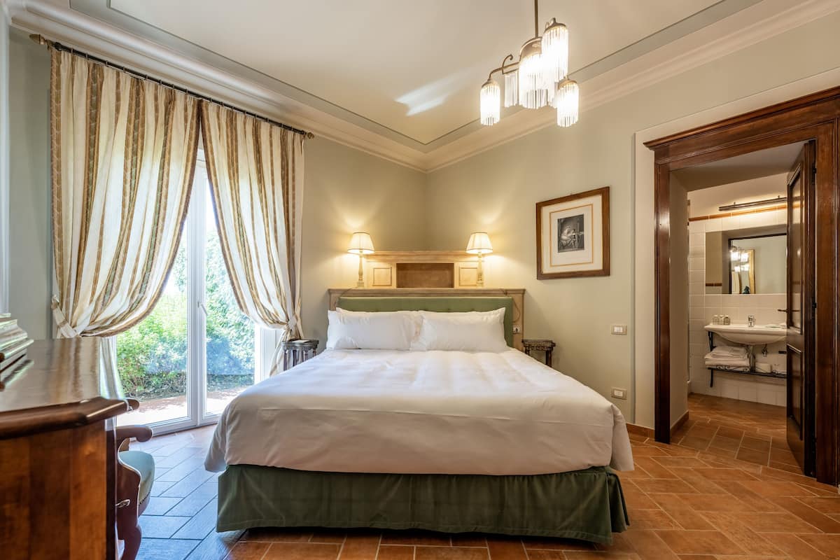 Een slaapkamer met een groot bed, twee bedlampen, een groen hoofdeinde, een houten bureau en uitzicht op het groen door een raam met gordijnen.