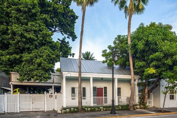 A white colonial-style building with a red door, surrounded by a white picket fence and palm trees.