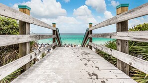 Ein hölzerner Steg führt zu einem Strand mit klarem türkisblauem Wasser und blauem Himmel.
