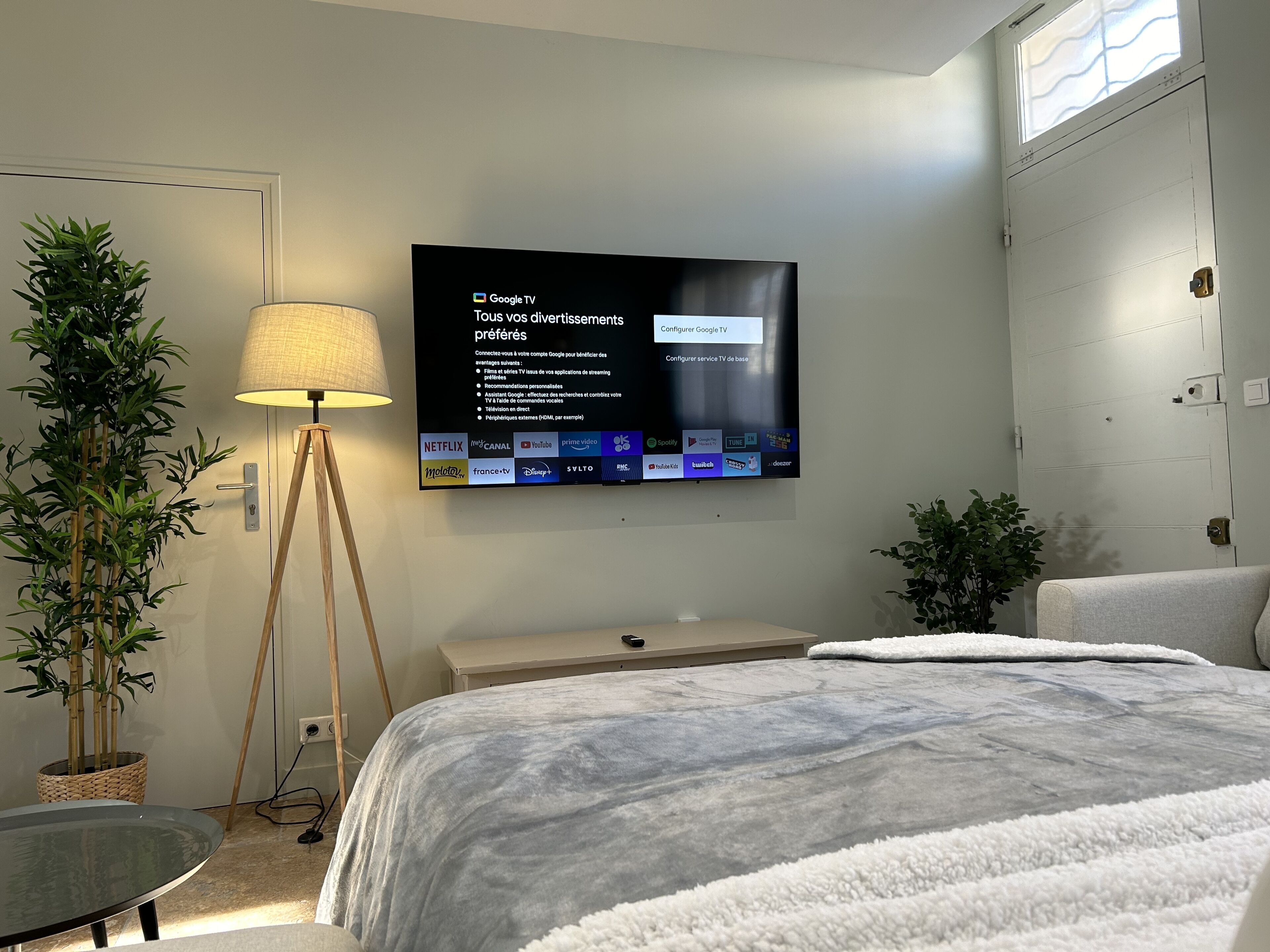 A hotel room with a bed, a TV mounted on the wall displaying Google TV interface, a lamp, and two potted plants.