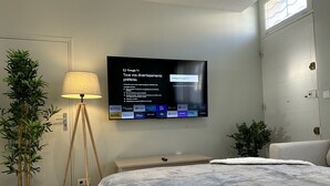Ein Hotelzimmer mit einem Bett, einem an der Wand befestigten Fernseher, der die Google TV-Oberfläche anzeigt, einer Lampe und zwei Zimmerpflanzen.