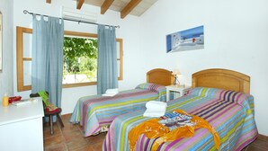 Een kamer met twee bedden, een bureau en een stoel. Er is een raam met gordijnen, een schilderij aan de muur en uitzicht op groen buiten.