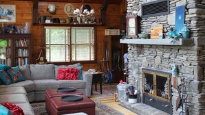 Un salon chaleureux avec une cheminée en pierre, des poutres en bois, une grande fenêtre et un ventilateur de plafond.