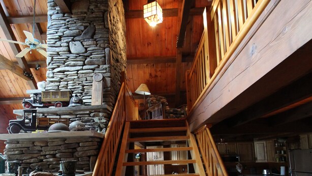 Un escalier en bois, avec un mur en pierre d’un côté et une rampe en bois de l’autre, mène à un espace situé au deuxième étage, où se trouvent des meubles de cuisine et un luminaire suspendu.