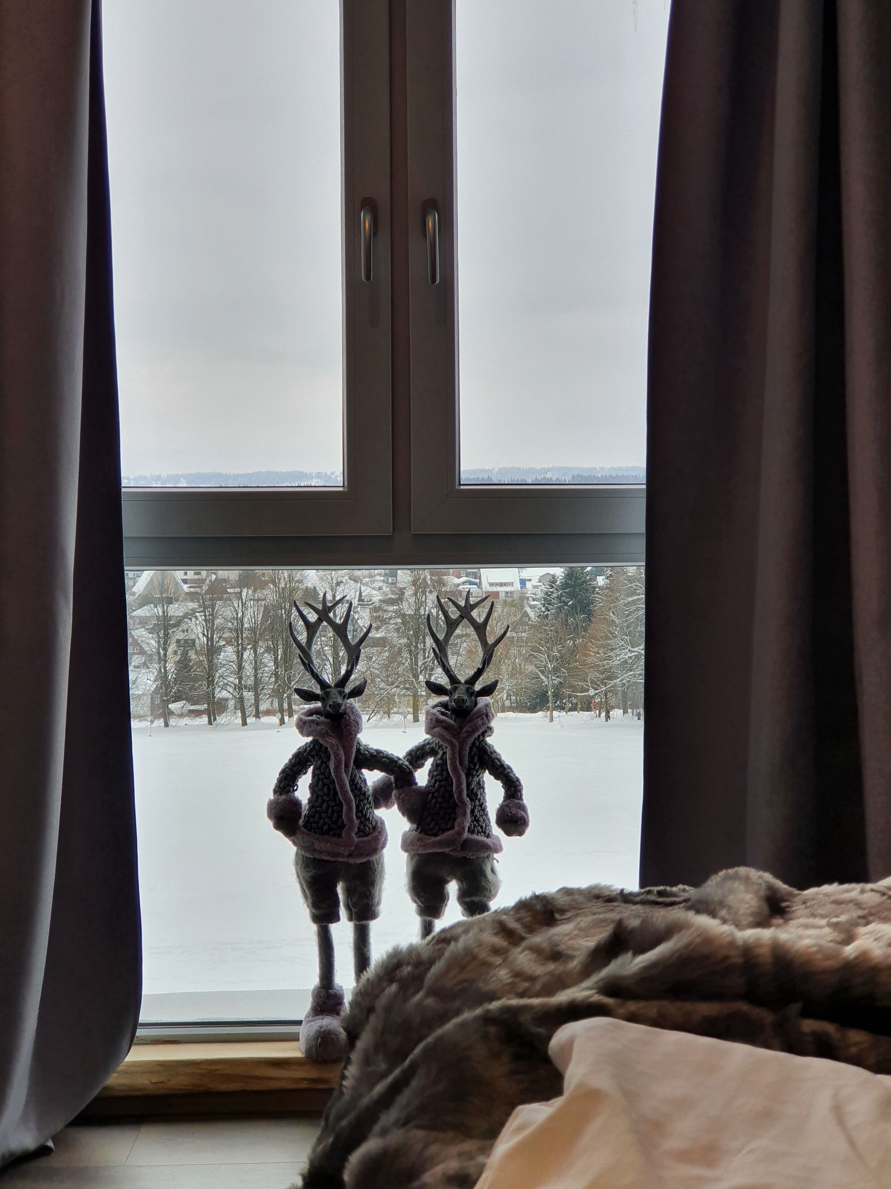 A window with two deer figurines on a sill, a view of a snowy landscape, and curtains partially drawn.