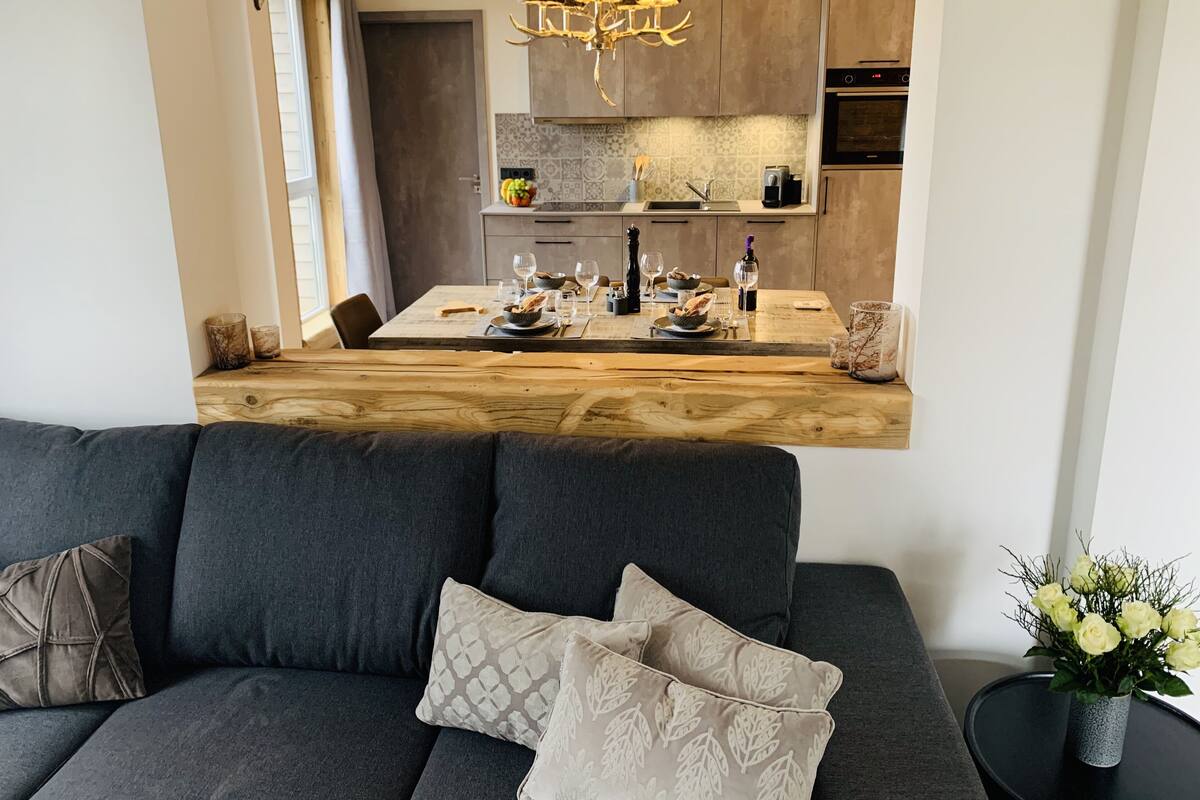 A modern kitchen with a central island, a dining area with a wooden table, and a dark grey sofa with patterned cushions.