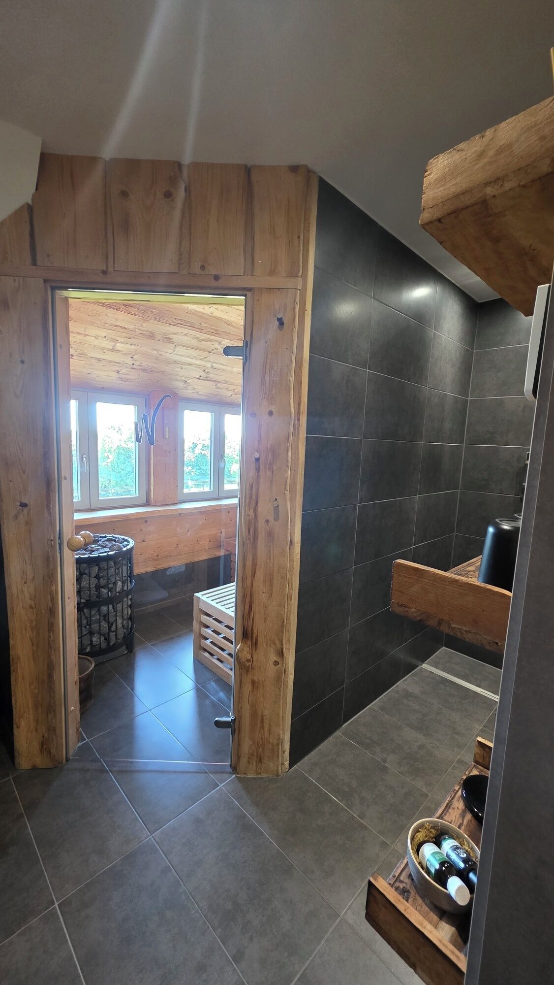 A modern bathroom with dark tiles, wooden accents, and a view of the outdoors.