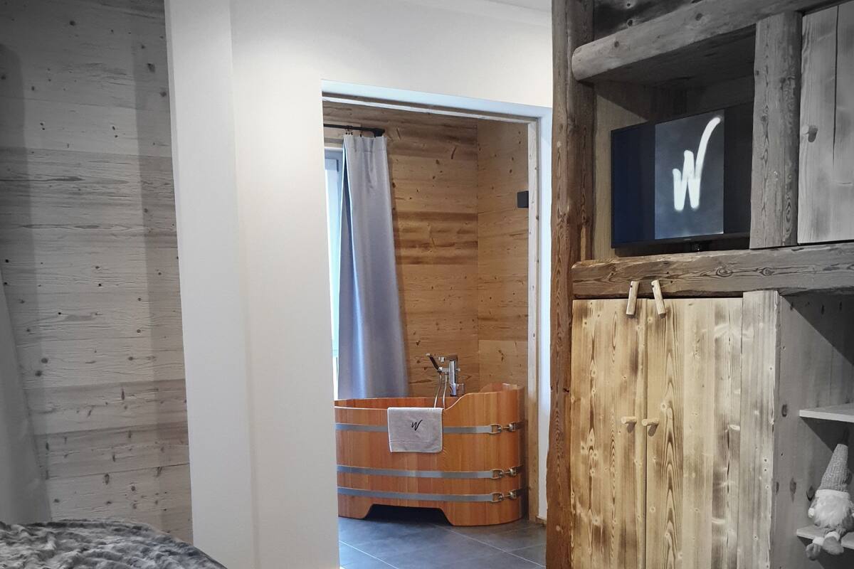 A bedroom with a bed, a wooden wardrobe, a small bathroom with a shower curtain, and a mounted TV.