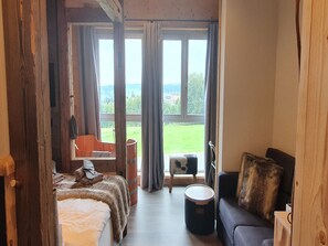 A room with a bed, a sofa, a chair, and a drum. There are large windows with curtains, and a view of a green landscape. - WLodgeOne (Braunlage)