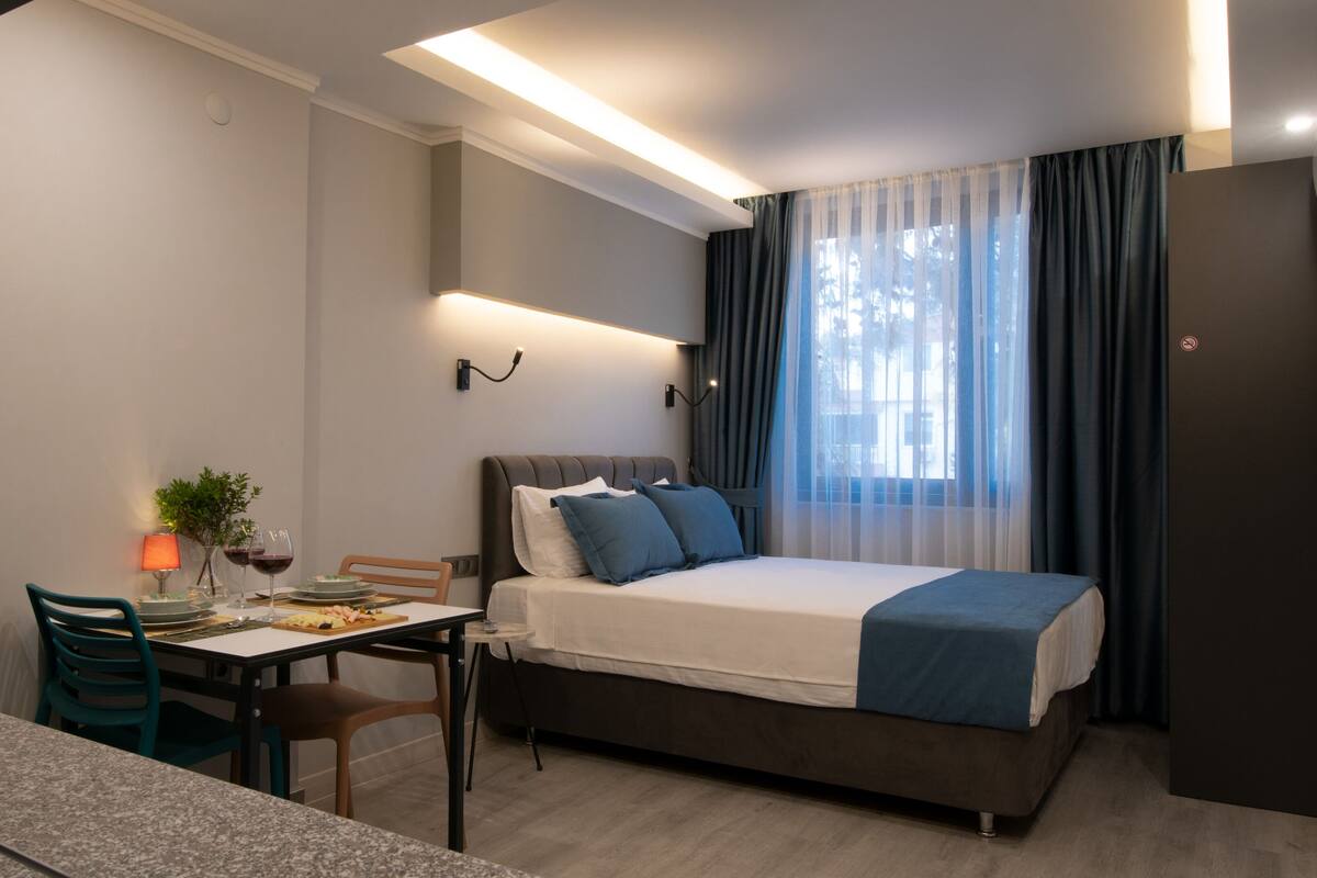 Una habitación de hotel moderna con cama, una mesa de comedor con sillas, una mesita con una planta y copas de vino, y vistas al exterior a través de cortinas translúcidas.