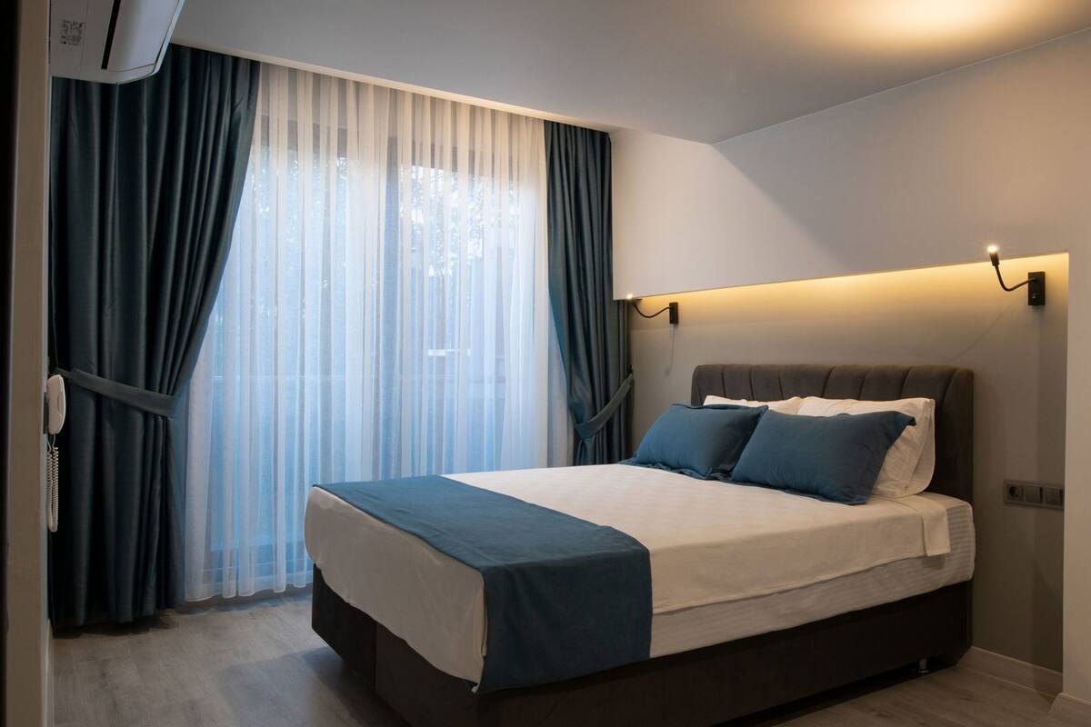 Una habitación de hotel moderna con una cama grande, cortinas oscuras, una mesita de noche y una lámpara de pared.