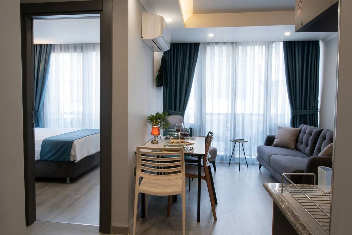 Una habitación de hotel moderna con cama, una mesa de comedor con sillas, un sofá y una pequeña mesa con una planta y una copa de vino.