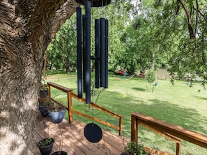A wooden deck with a hanging wind chime, surrounded by trees and a hammock in the background.