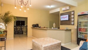A hotel reception area with a counter, a television displaying the hotel name, a refrigerator stocked with beverages, and a seating area with an ottoman. - Smart Hotel Rio Verde (Rio Verde)