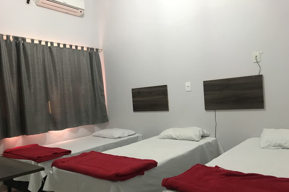Ein Hotelzimmer mit drei Einzelbetten, jedes mit einer weißen Matratze und einer roten Decke. Ein kleiner Tisch mit einer roten Decke, und ein grauer Vorhang ist teilweise heruntergezogen.
