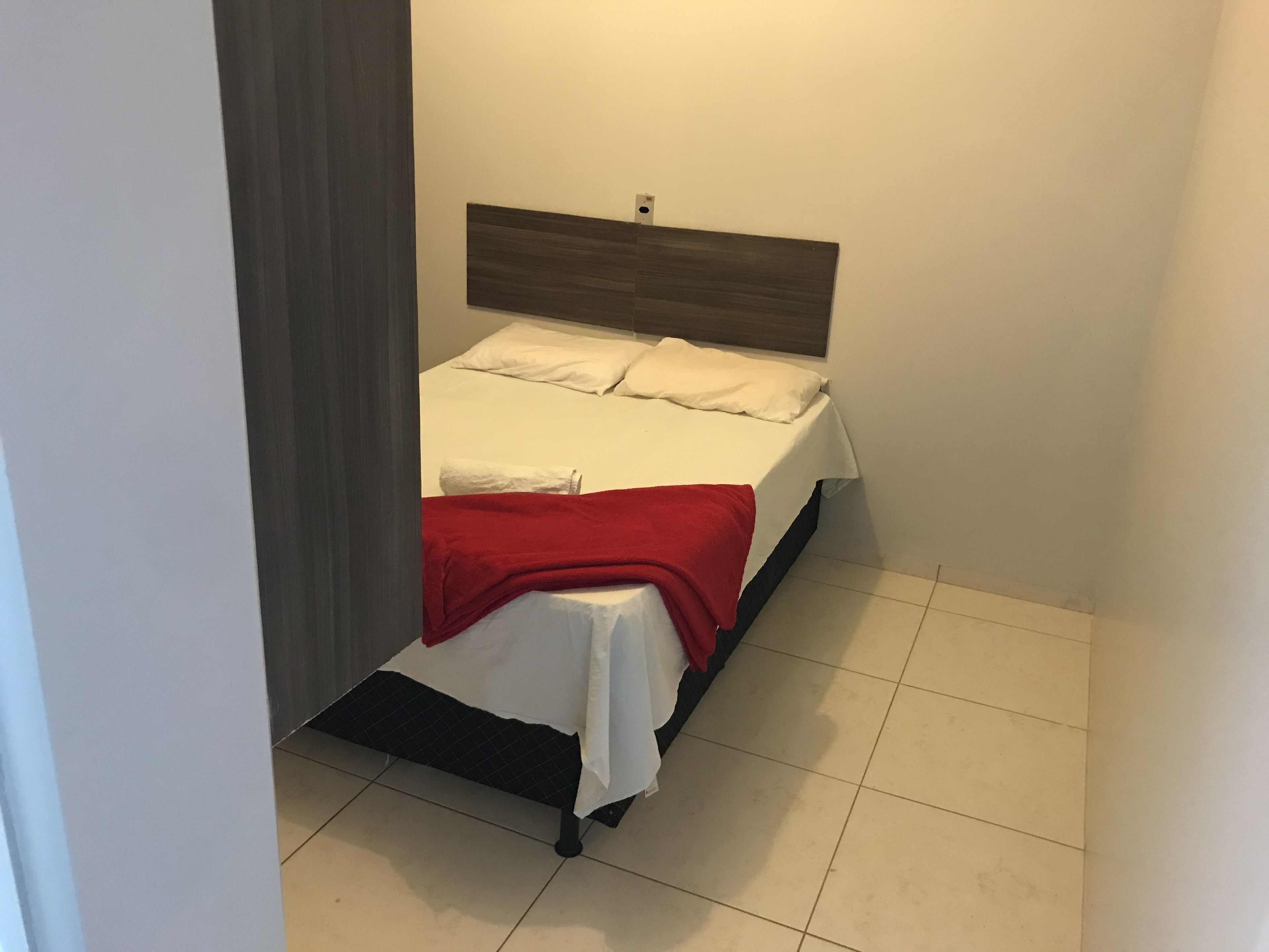 A small, neatly made bed with white linens and a red blanket, positioned in a corner room with white walls and a tiled floor.