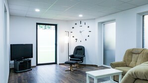 Una sala de estar moderna con un televisor de pantalla plana, una silla de oficina negra, un sofá color beige y una mesa de centro blanca.