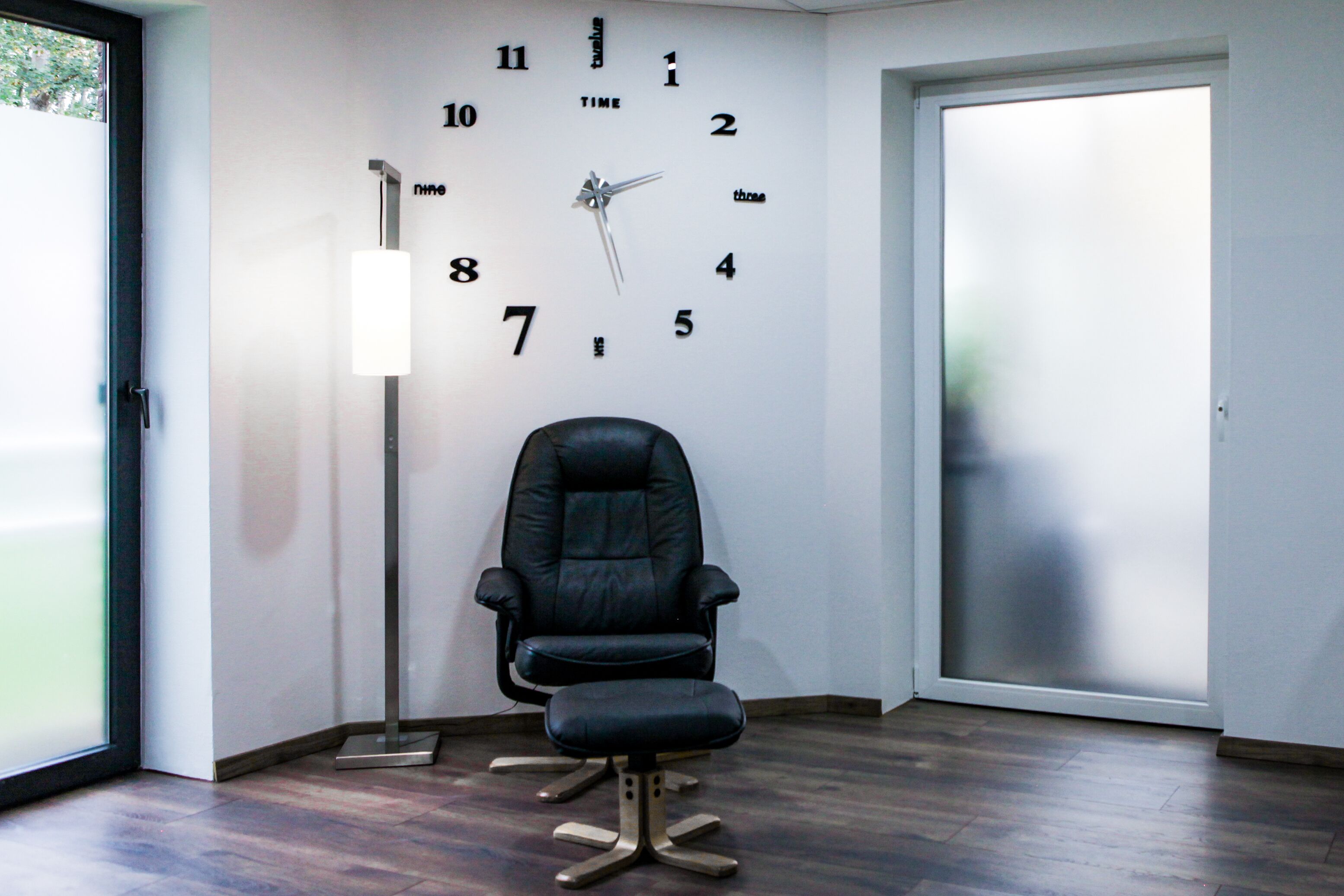 Ein modernes Zimmer mit schwarzem Bürostuhl, einer Wanduhr und einer Stehlampe.