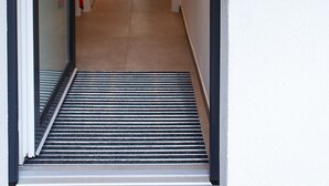 Un pasillo estrecho con un umbral metálico, una puerta de vidrio y una lámpara fijada a la pared.