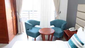 A hotel room with two blue armchairs, a wooden table, a bed with white bedding, a bedside table with a phone, and a view through sheer curtains. - Meridian C.A (Dar es Salaam)