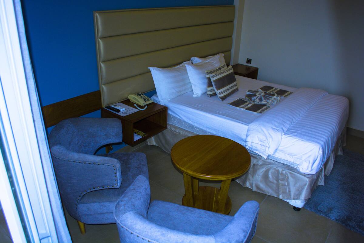 A hotel room with a bed, two armchairs, a small wooden table, a nightstand with a telephone, and a wall-mounted shelf.