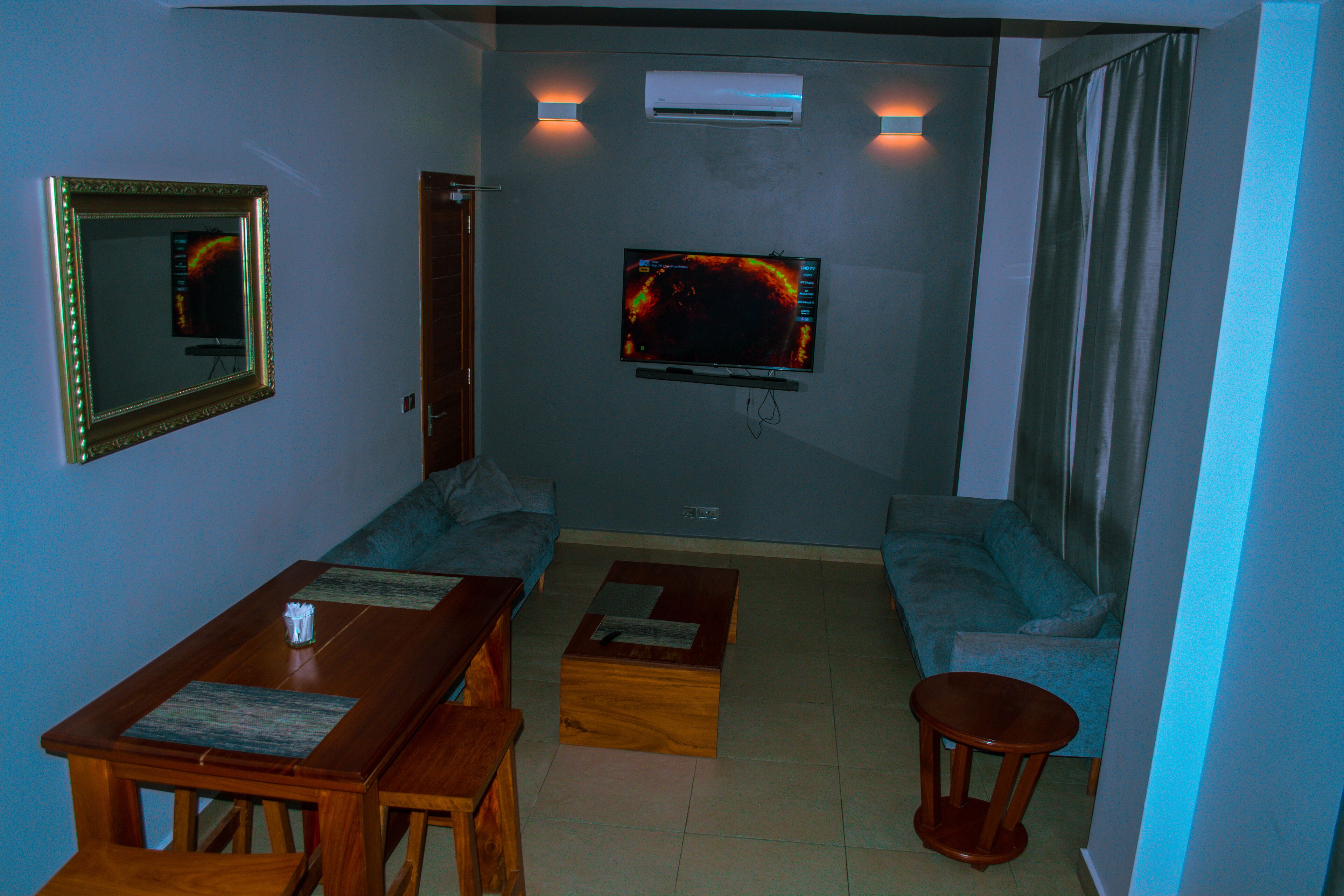A living room with a wooden dining table, two sofas, a TV mounted on the wall, and an air conditioning unit.
