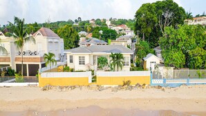 Une propriété située en bord de mer, offrant une vue dégagée sur l’océan, une plage de sable et un quartier résidentiel avec des maisons et de la verdure en arrière-plan.