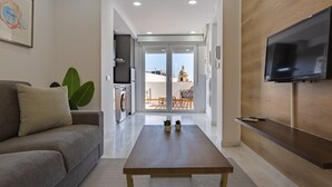A modern living room with a sofa, a coffee table, a TV mounted on the wall, and a view of the balcony and outside.