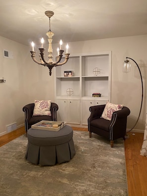 Un salon avec un lustre, deux fauteuils, un pouf rond et une lampe sur pied.