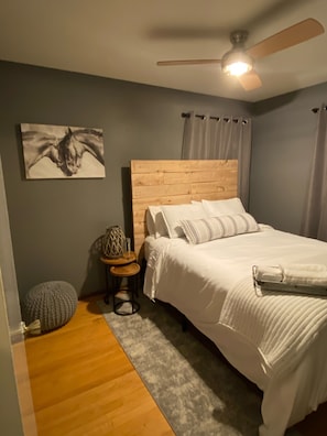 Une chambre à coucher avec une tête de lit en bois, un lit recouvert de linge de lit blanc, un tapis gris, un petit pouf rond, une table de chevet avec une lampe et un ventilateur de plafond.