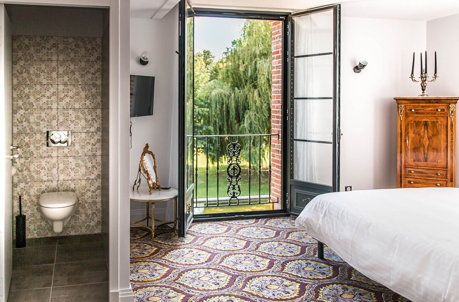Une chambre à coucher avec un lit, une armoire en bois, une salle de bain avec des toilettes et un balcon donnant sur un espace verdoyant.
