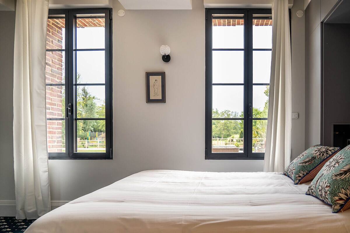 A bedroom with a large bed, two windows with curtains, a framed picture on the wall, and a view of the outside.
