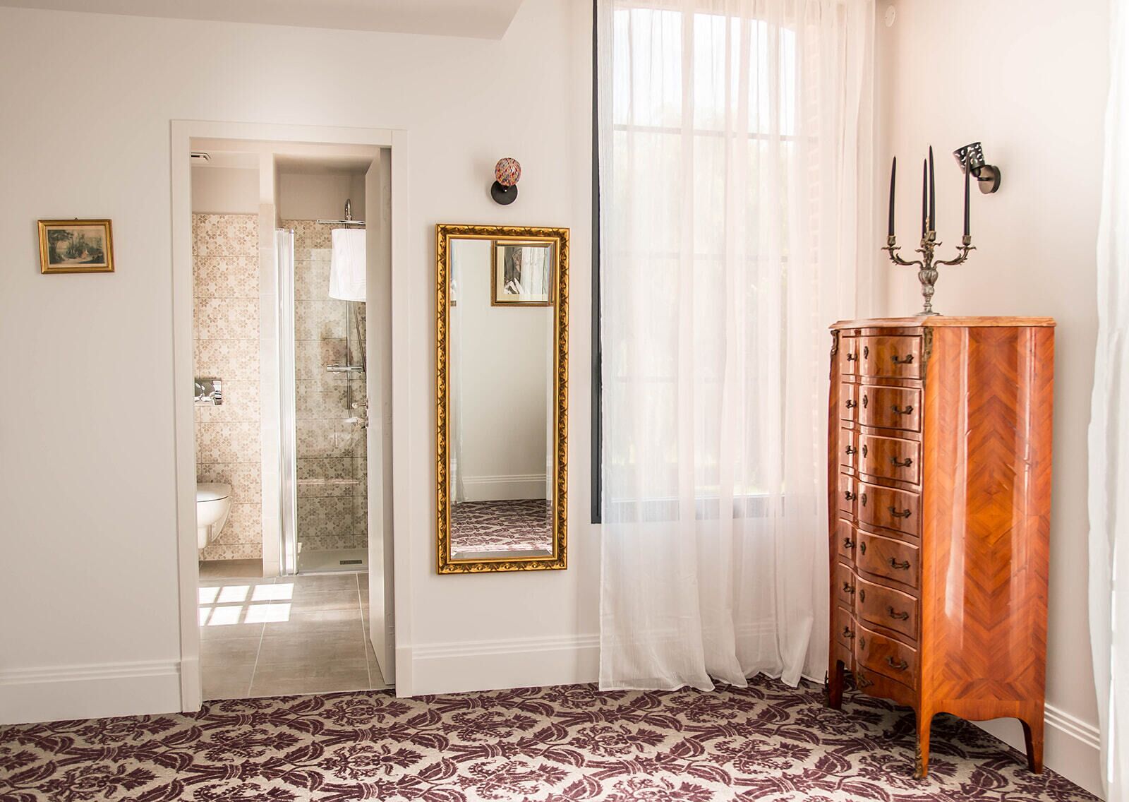 Une pièce dotée d’un tapis à motifs, d’une commode en bois avec des tiroirs, d’un miroir avec un cadre doré et d’une porte donnant sur une salle de bain équipée d’une douche et de toilettes.
