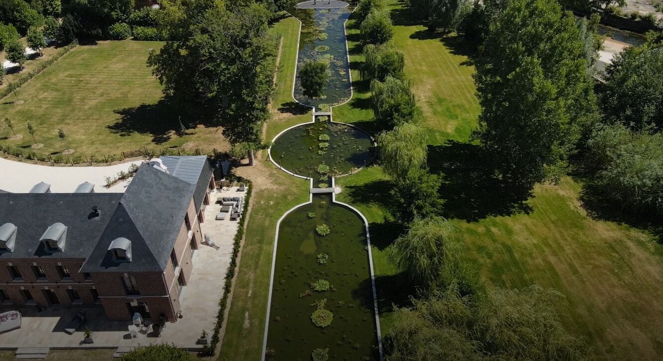 Luchtbeeld van een aangelegd gebied met een centraal waterpartij, omringd door bomen en een gebouw met een schuin dak.