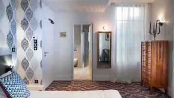 Un dormitorio con papel tapiz estampado, una cama con cubrecama blanco, un aparador de madera, un espejo y una araña.