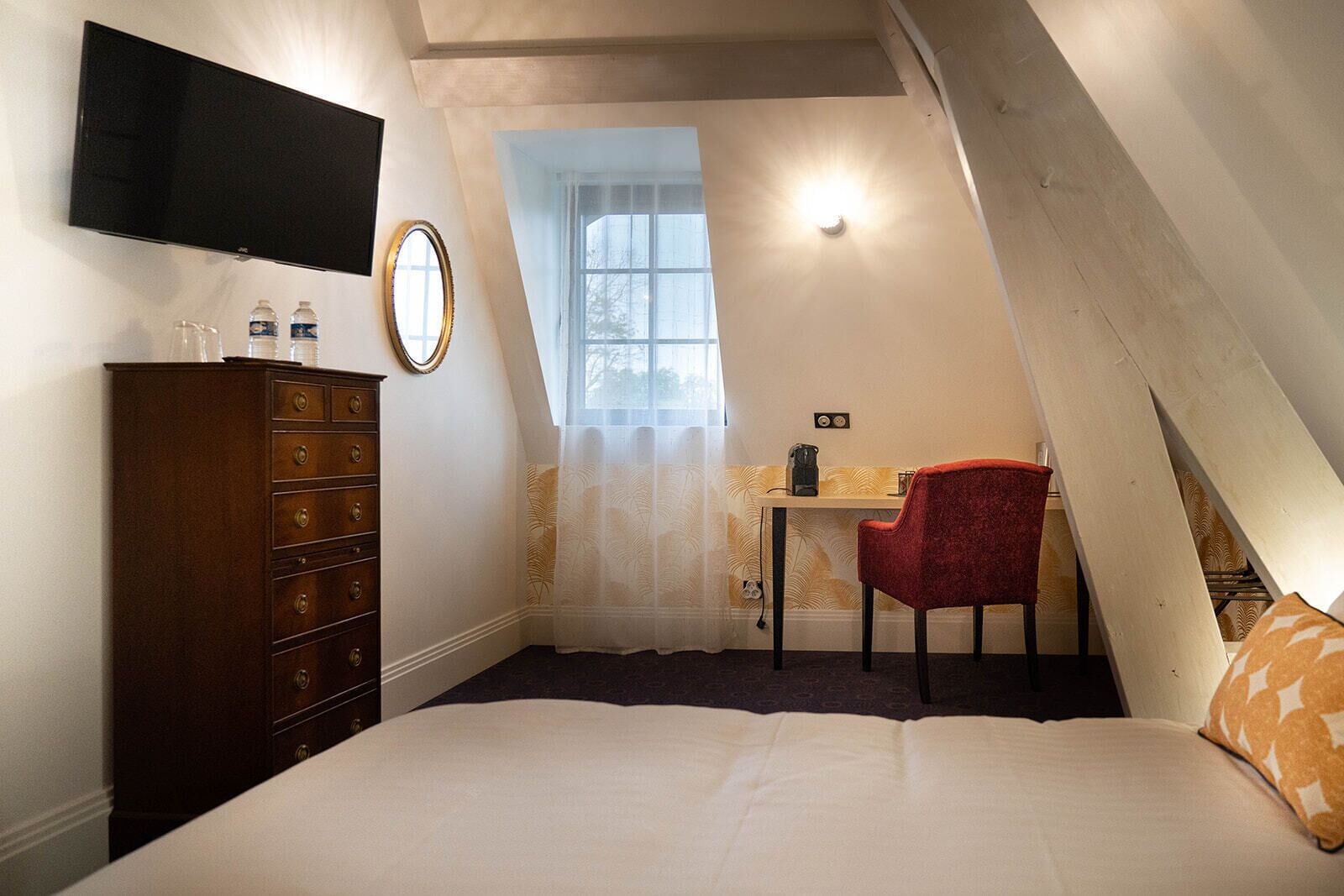 A bedroom with a bed, a dresser, a desk with a chair, a TV mounted on the wall, and a window with a curtain.