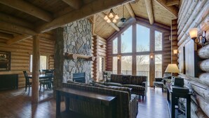 A log cabin interior with a stone fireplace, wooden furniture, and large windows.