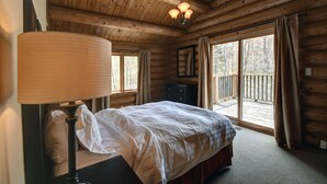 A wooden cabin bedroom with a bed, a nightstand, a lamp, a dresser, and a large window with curtains.