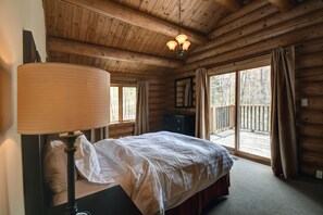 A wooden cabin bedroom with a bed, a nightstand, a lamp, a dresser, and a large window with curtains.