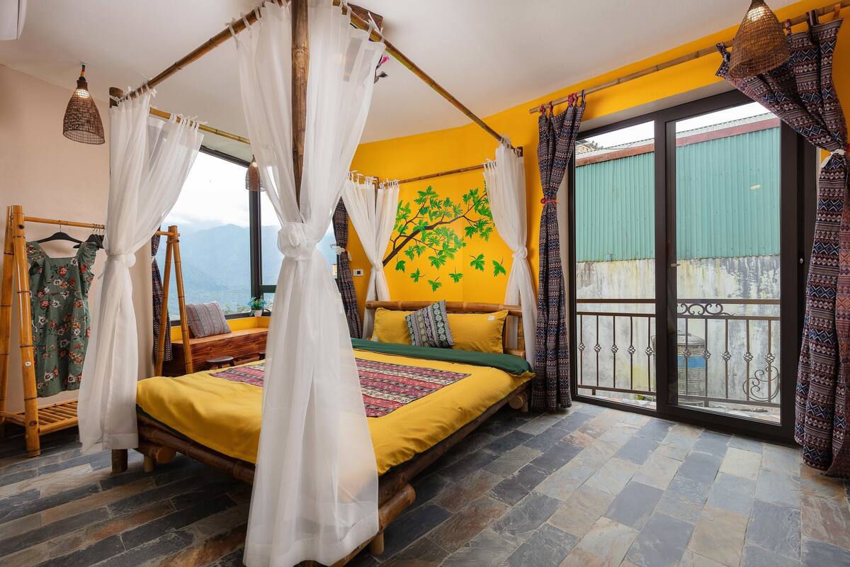 A room with a bed, a window with curtains, a view of mountains, and a balcony with a railing.