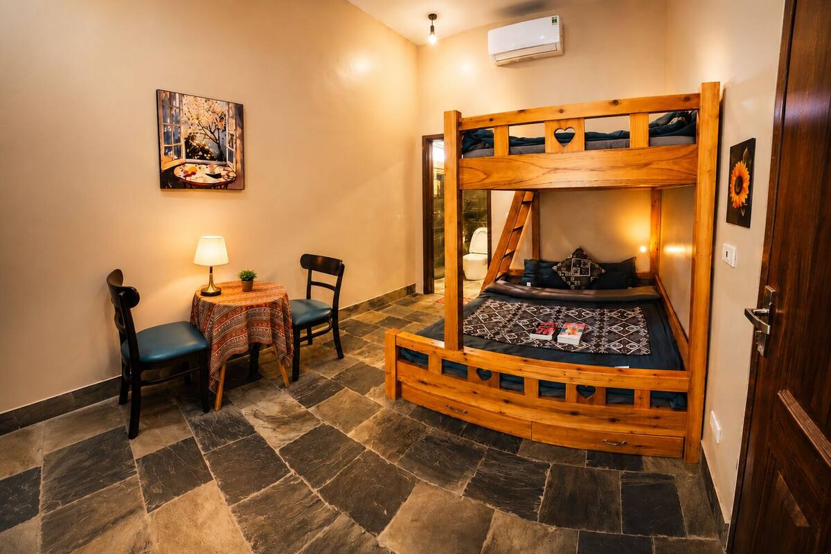 A bunk bed with a curved wooden frame, a small table with a lamp, two chairs, and a painting on the wall.