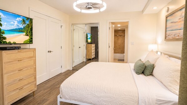 Un dormitorio con cama, televisor, cómoda y un baño visible a través de una puerta abierta.