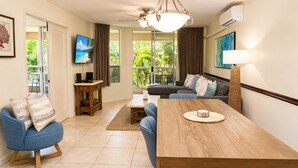 A modern living room with a sofa, a dining table, a TV, and a view of greenery outside. - Rare 3 Bedroom Penthouse at Maui Banyan | All En Suites and Heart of Kihei (Kihei)