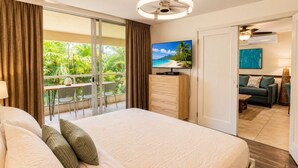 A hotel room with a bed, a TV, a wooden dresser, a balcony with chairs, and a view of greenery. - Rare 3 Bedroom Penthouse at Maui Banyan | All En Suites and Heart of Kihei (Kihei)