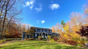 A two-story house with a front porch, surrounded by a well-maintained lawn and autumn-colored trees. - Hidden Oasis in the Mountains by Evergreen Home (Pittsfield)
