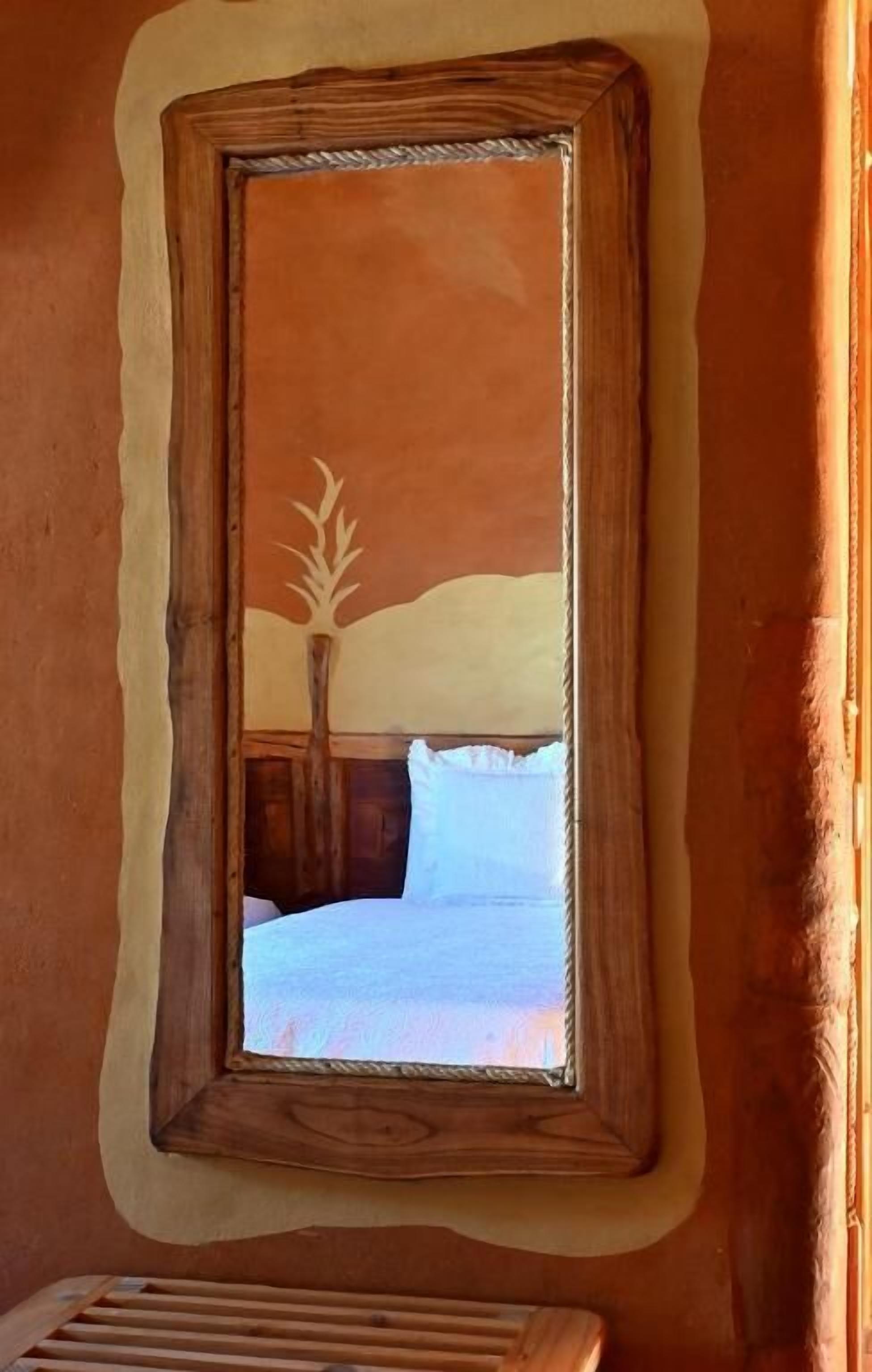 A room with a wooden-framed mirror, a bed with white bedding, and a window with a view of a tree.