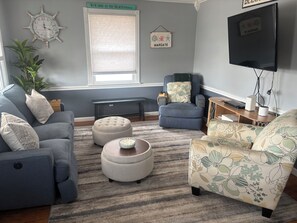A living room with a grey sofa, blue armchair, ottoman, and floral-patterned chair. There's a clock, a sign reading 'Welcome to the Beach House,' and a television on a stand.